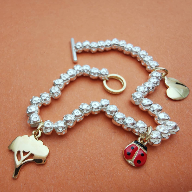 Bracciale artigianale in argento 925 con ciondoli dorati a forma di albero e cuore e una coccinella smaltata rossa, realizzato a mano da ViMon Gioielli su sfondo arancione