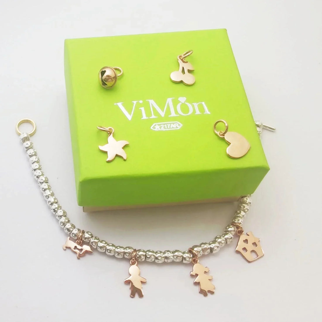 Bracciale con charm in oro rosa giallo e perle argentate ViMon, con ciondoli a forma di stella, bambina, albero e casetta, accanto a scatola verde elegante con logo ViMon