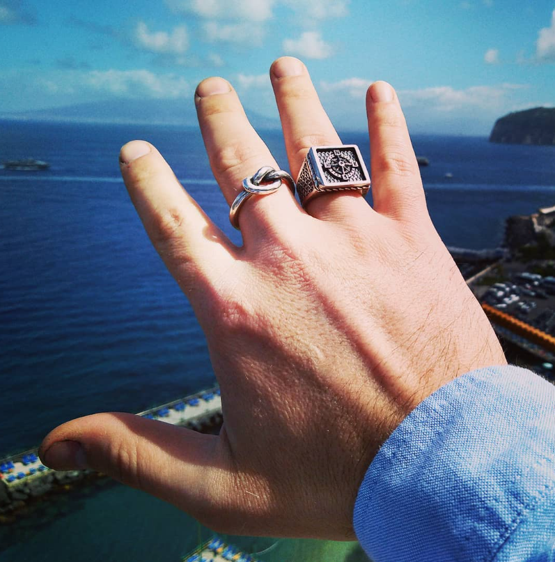 Mano con due anelli artigianali in argento 925, uno chevalier inciso e uno ad anello nautico, indossati con vista sul mare, realizzati da ViMon Gioielli.