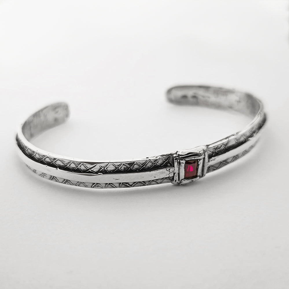 Bracciale rigido da uomo in argento 925 artigianale con incisioni geometriche e pietra rossa quadrata centrale, fotografato in primo piano su fondo bianco minimal.