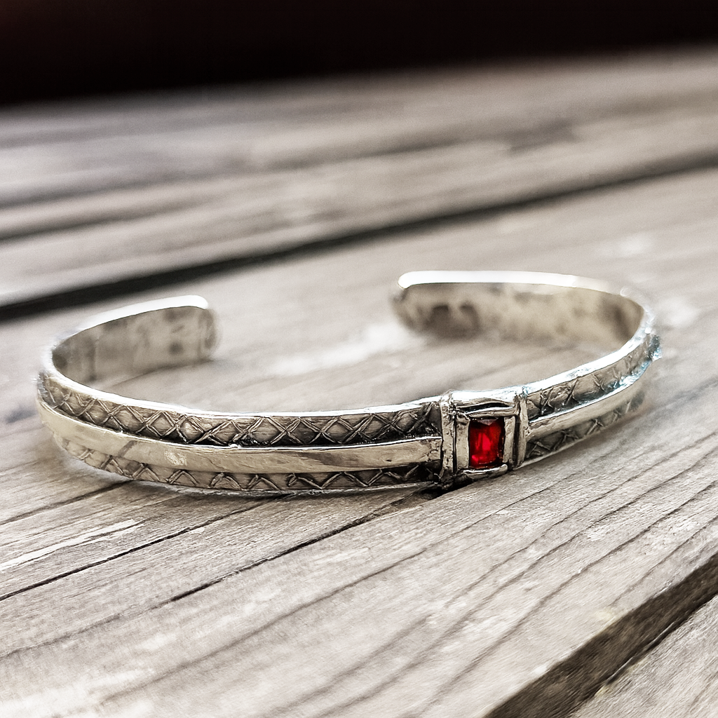 Bracciale rigido da uomo in argento 925 con incisioni geometriche e pietra corindone rosso taglio quadrato, appoggiato su superficie in legno.