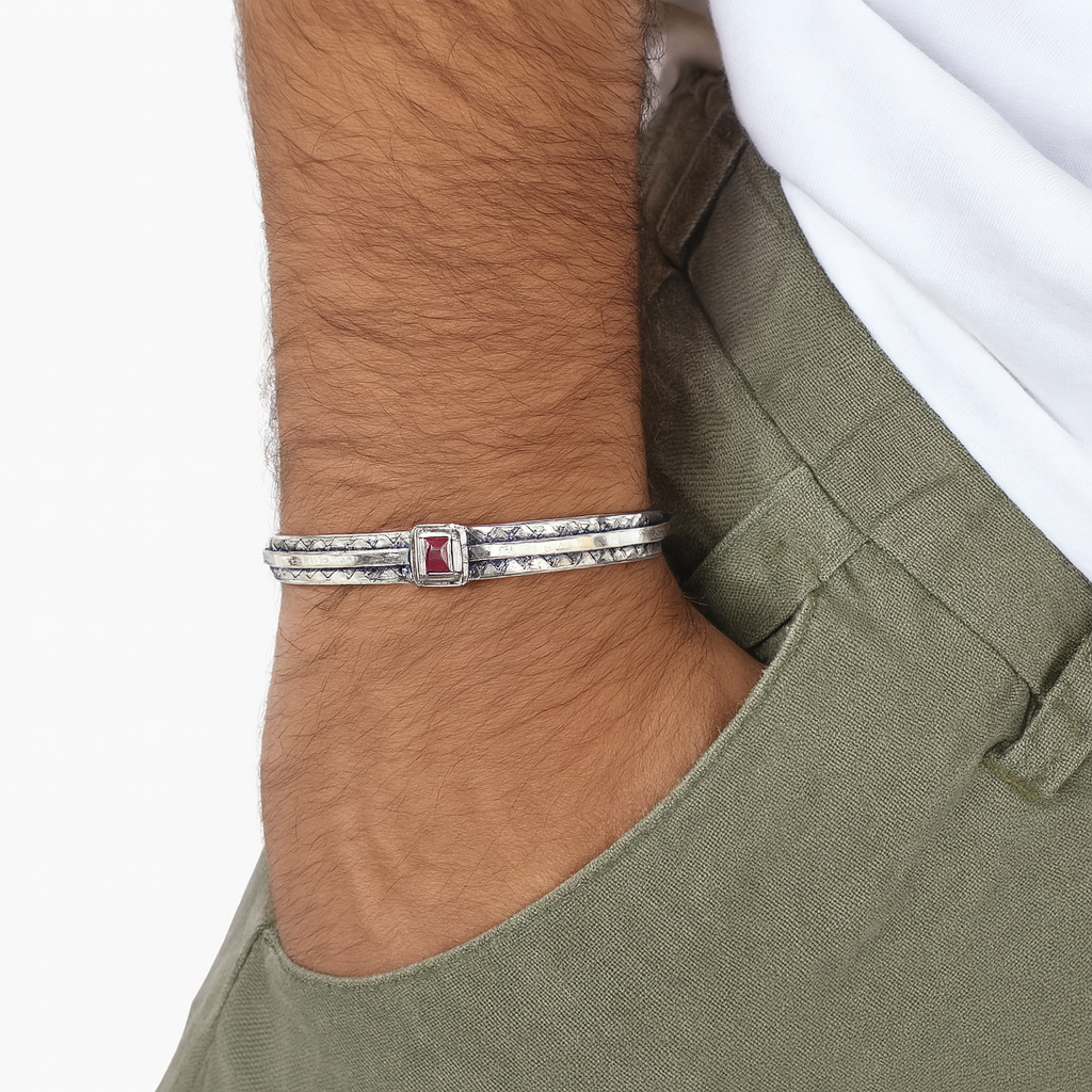 Bracciale rigido da uomo in argento 925 con incisioni geometriche e pietra corindone rosso taglio quadrato, indossato al polso sopra pantaloni verde oliva e maglietta bianca, su sfondo bianco neutro.